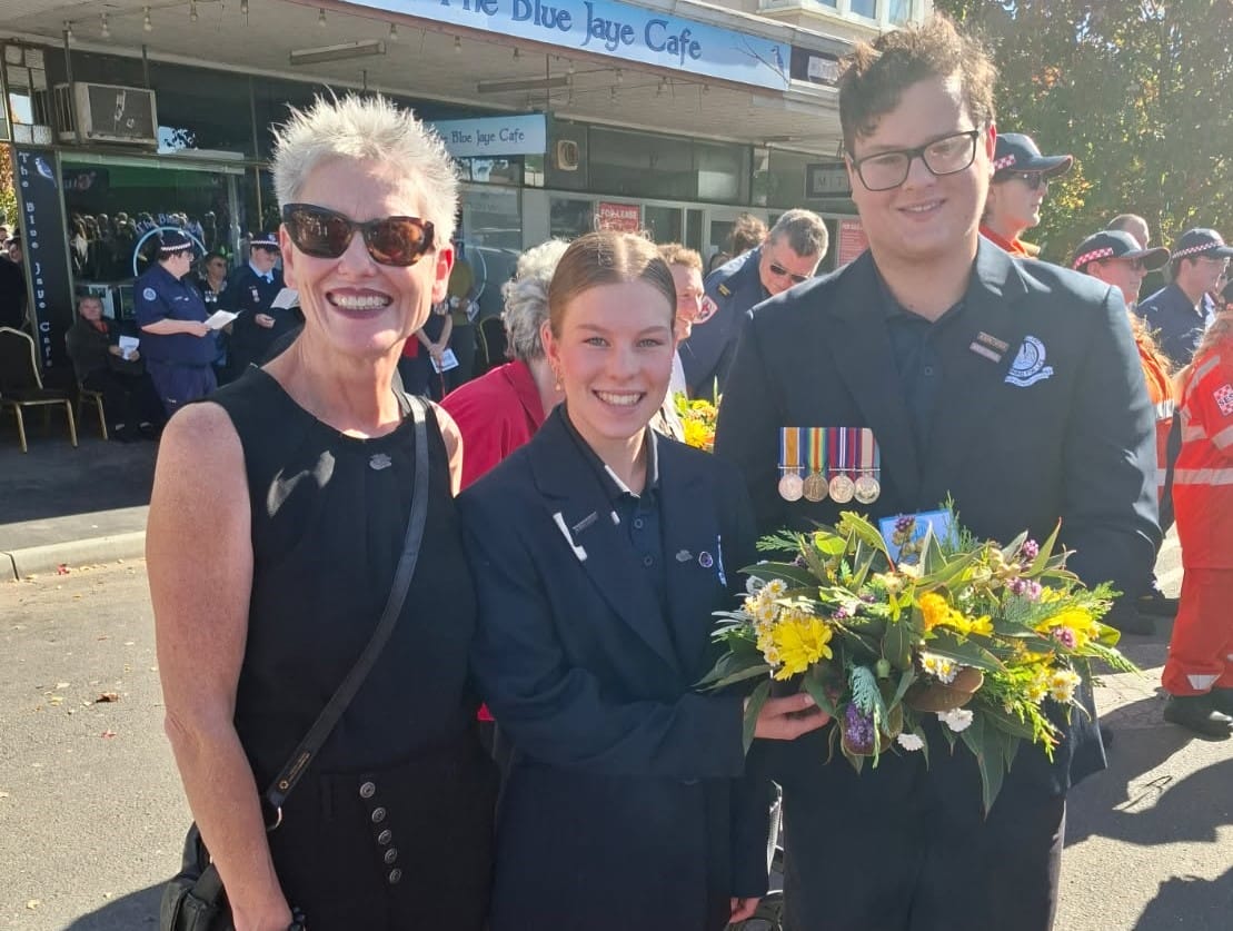 Smiling trio posing at a parade: a woman in black, a young woman in a navy blazer, and a medal-winning young man holding a bouquet outside a cafe.