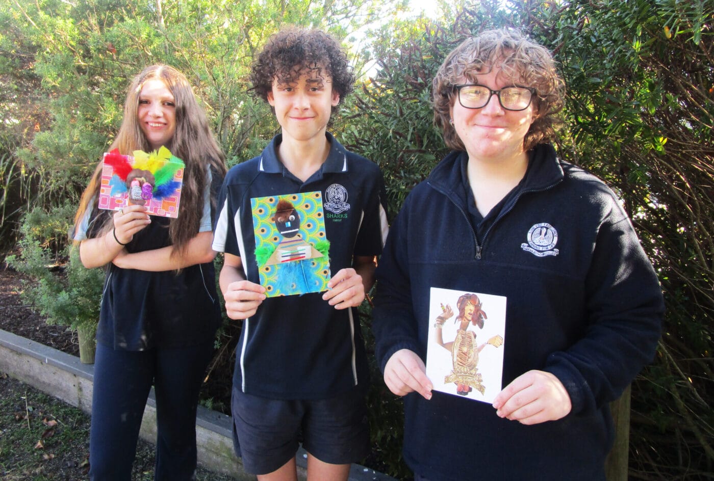 Three teenagers outdoors proudly displaying handmade crafts and cards they've made, standing in front of green bushes.