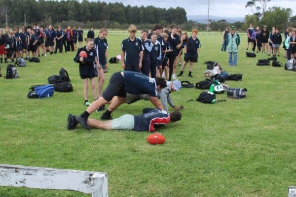 Young rugby players on a grassy field watching as one player tackles another near a red rugby ball amidst a group of teammates and backpacks in the background.