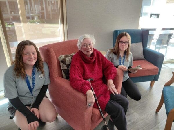 Smiling elderly woman in a red sweater sits on a peach sofa with a cane, flanked by two young caregivers in gray shirts in a bright lounge.