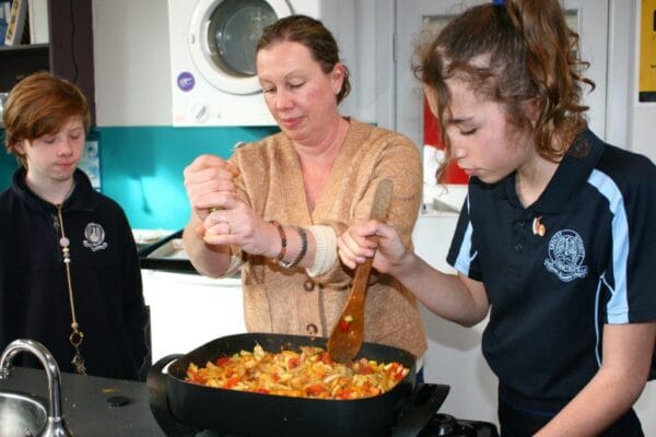 Woman instructor supervising two students as they stir a vegetable stir-fry in a large skillet on a stove, in a bright kitchen.