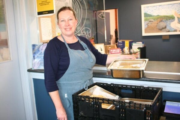 Smiling woman in a blue apron stands behind a cafe counter with metal trays of food.