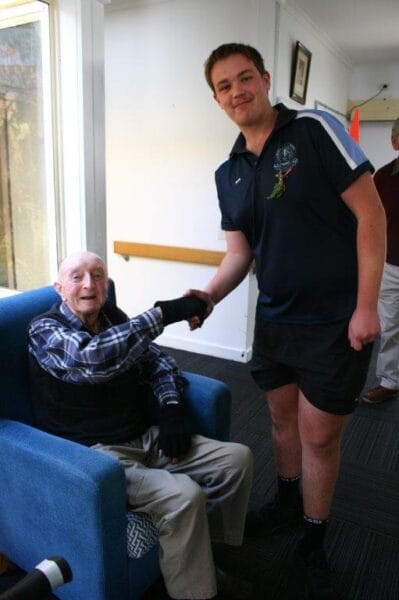 A young man in a navy polo shirt shakes hands with an elderly man in a plaid shirt, who is seated in a blue armchair by a window in a bright room.