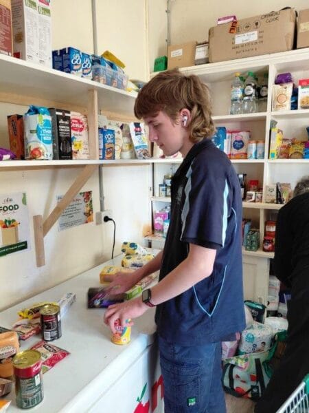 A young person in a navy shirt sorting canned goods on a pantry counter with shelves of food behind them, wearing a wireless earbud.