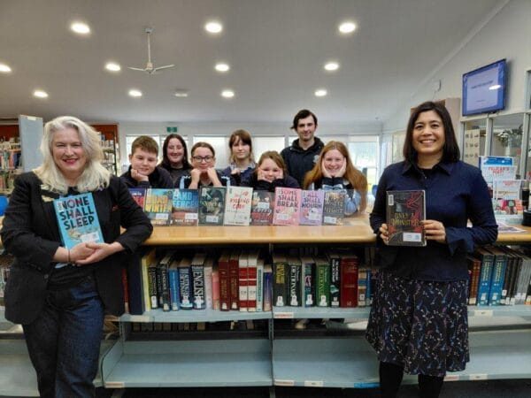 Group of people of varying ages in a library, smiling and posing behind a table of colorful books on display.