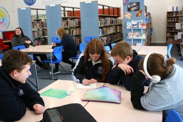 Group of four students studying at a library table, two writing and two discussing, with bookshelves in the background.