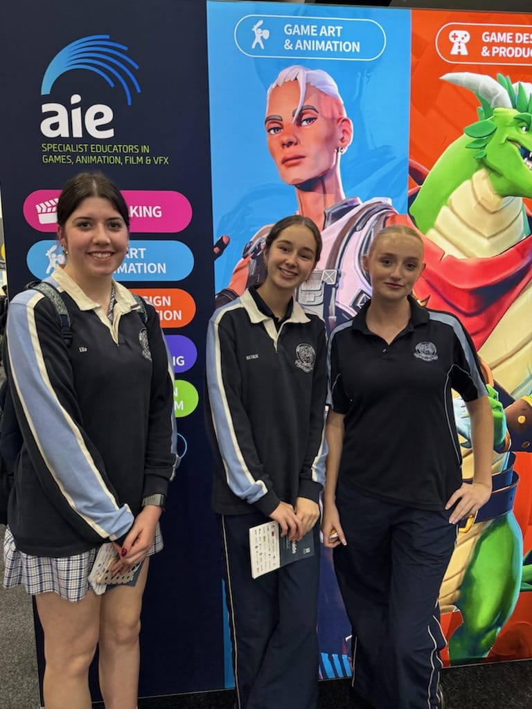 Three smiling female students in school uniforms pose in front of a colorful game art booth at a expo, with banners reading Game Art & Animation and related programs.