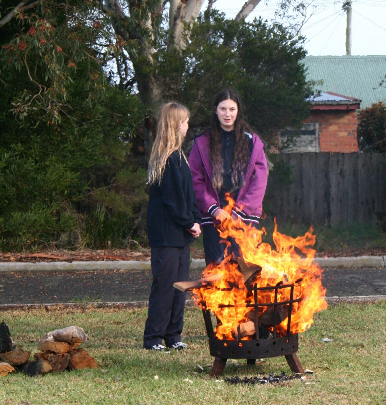 Two girls stand beside a large outdoor fire in a metal fire pit, burning logs, in a grassy yard with trees in the background.