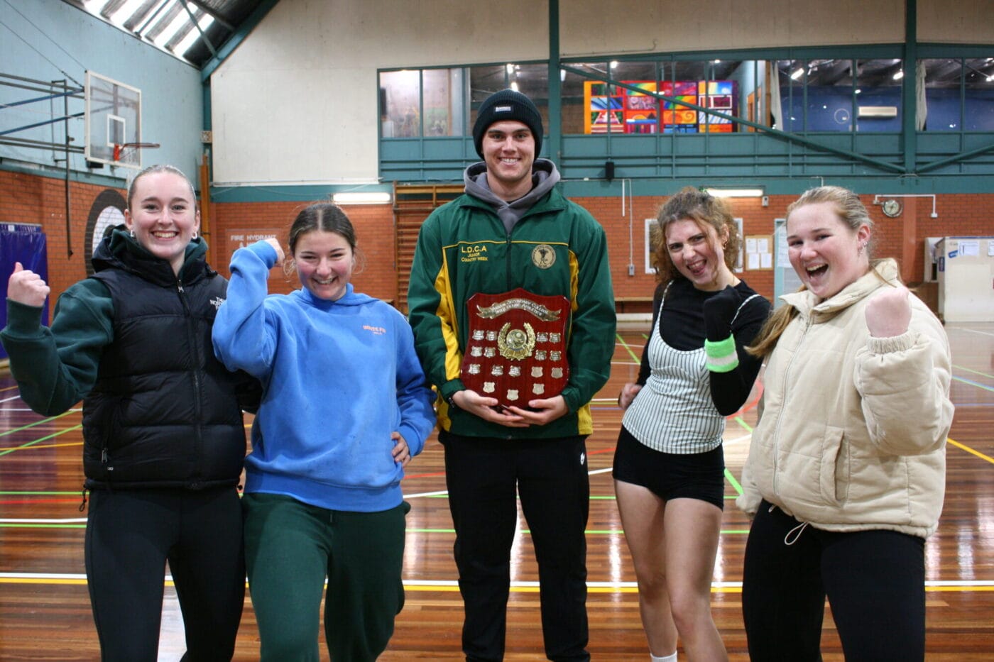 Five young adults in athletic wear pose in a gym, smiling with a large trophy held by the center person